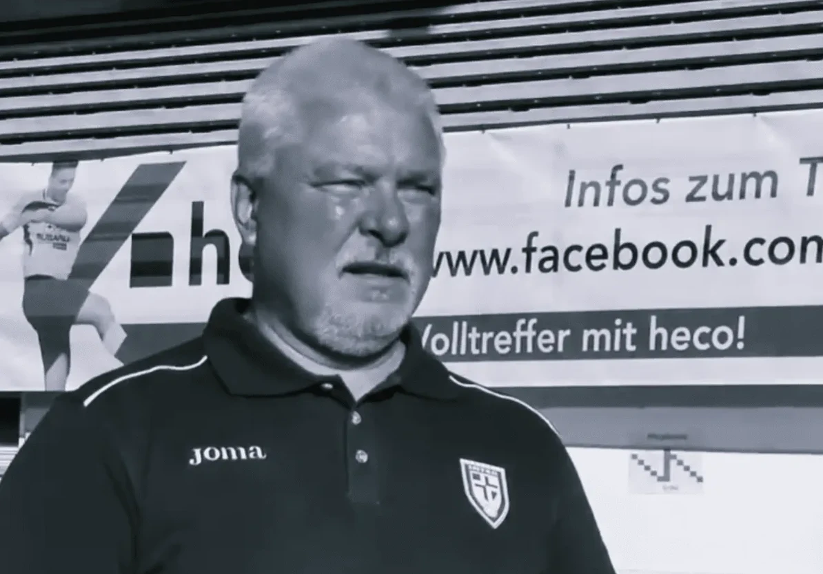 Muškarac sa sijedom kosom i bradom nosi crnu majicu sa amblemom.