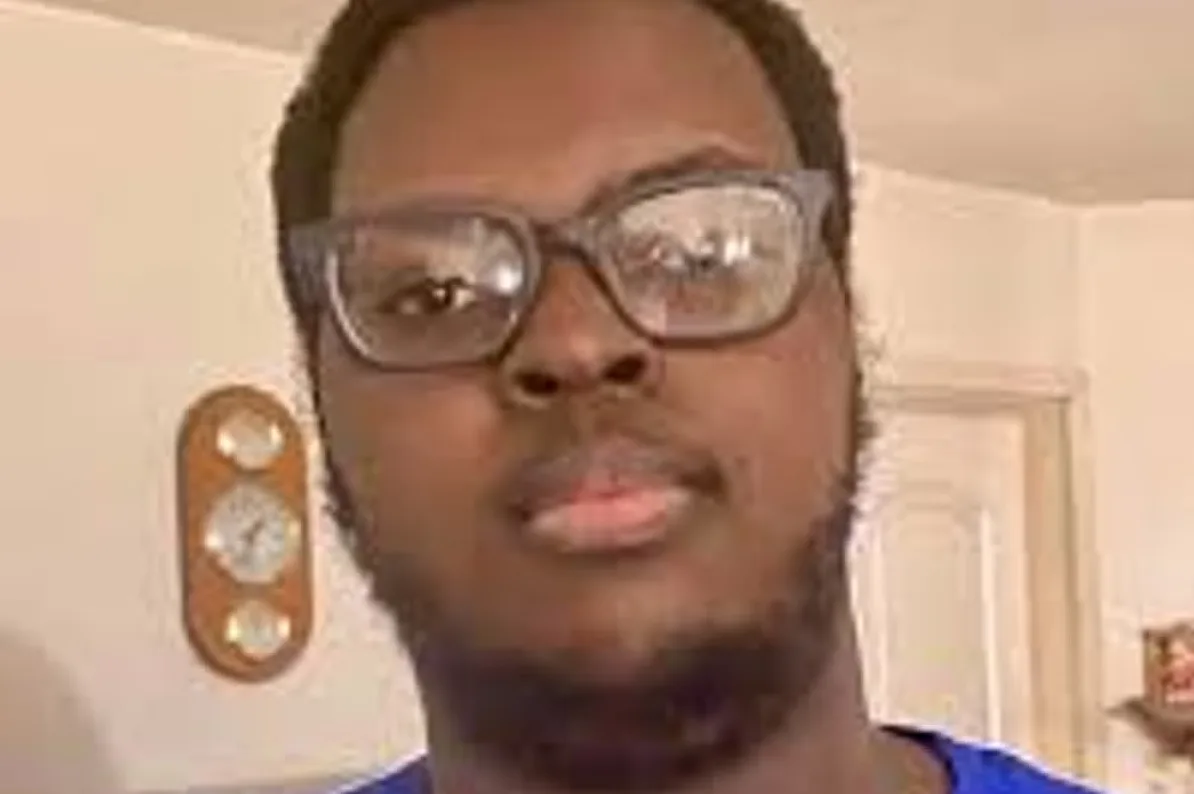 Crnac s naočalama i bradom pozira ispred sata na zidu.