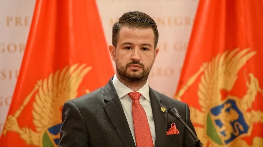 Portret crnogorskog političara sa bradom, obučenog u tamno odijelo i crvenu kravatu sa bijelim dezenom, te crvenom maramicom u džepu. U pozadini se vide zastave Crne Gore sa zlatnim dvoglavim orlom.