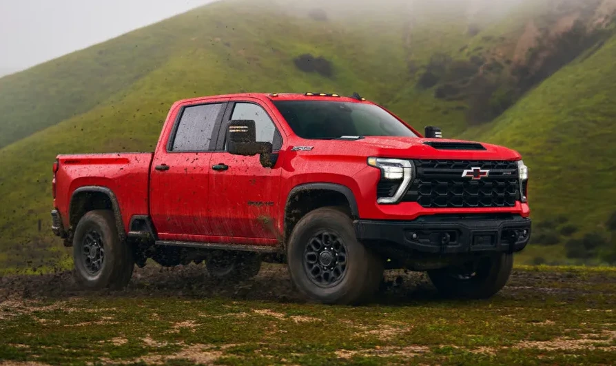 Crveni Chevrolet Silverado kamionet vozi po blatnjavom terenu sa zelenim brdima u pozadini.