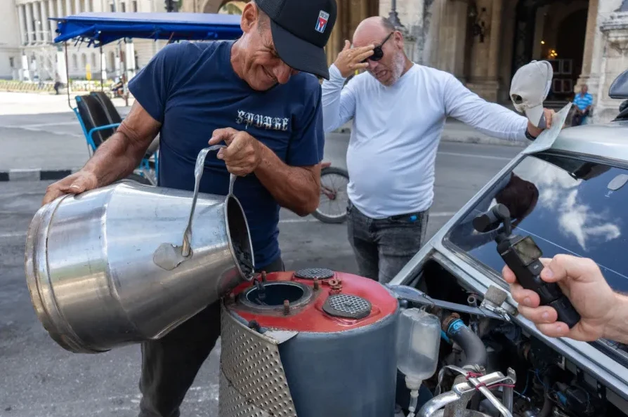Čovjek u plavoj majici sa crnim šeširom pretače gorivo iz metalnog kanistera u rezervoar starinskog automobila.