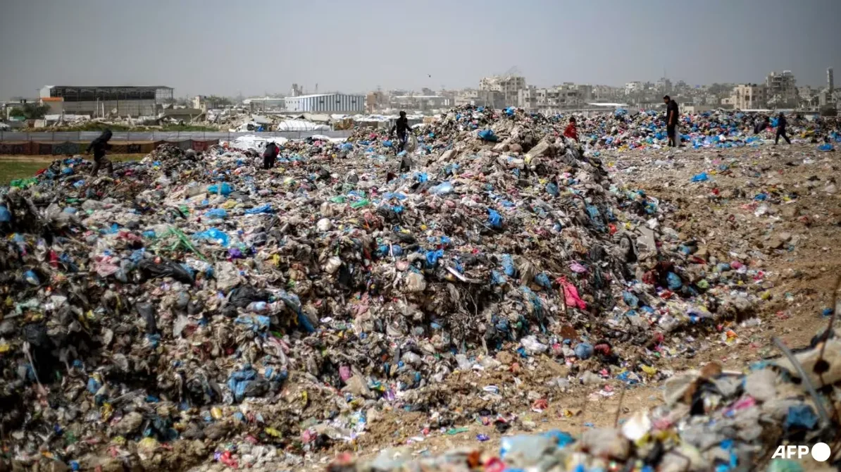 Ljudi, uključujući djecu, pretražuju ogromnu deponiju otpada pod vedrim nebom, sa zgradama u daljini.