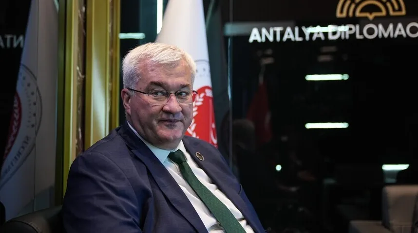 Muškarac u tamnom odijelu i bijeloj košulji sa zelenom kravatom, nosi naočale i sjedi u fotelji pored zastava.