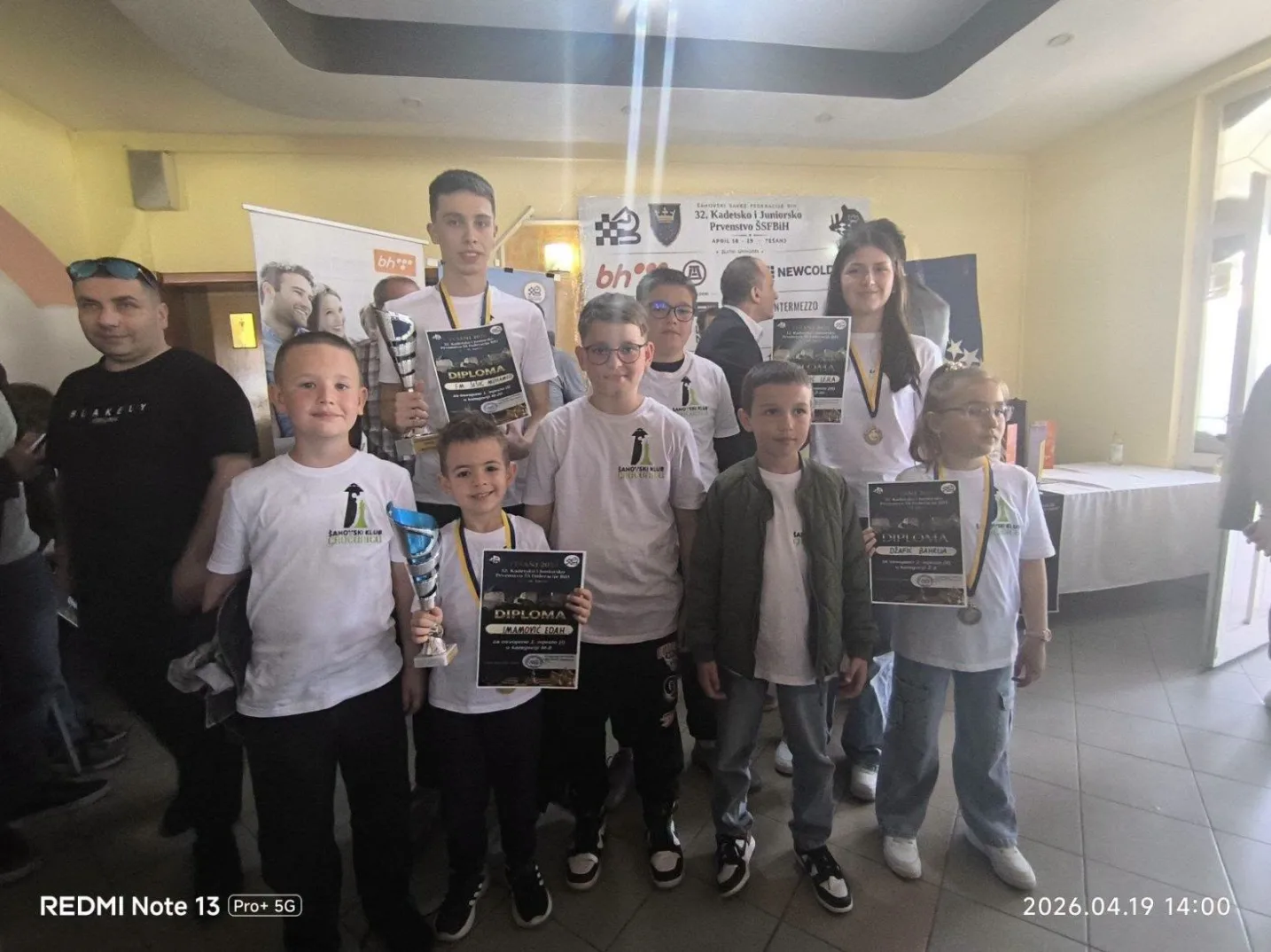 Grupa djece i odraslih pozira za fotografiju nakon šahovskog takmičenja, držeći diplome i pehare.