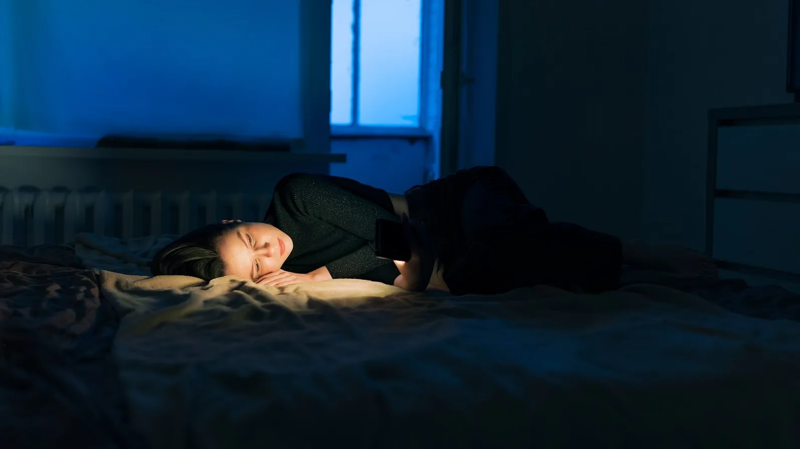 Mlada djevojka leži na krevetu u mraku i gleda u ekran svog pametnog telefona, koji osvjetljava njeno lice.