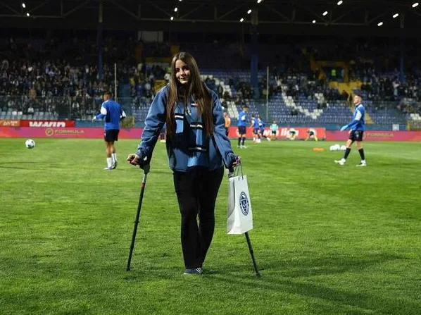 Djevojka sa štakom u ruci i torbom stoji na nogometnom terenu, s igračima u pozadini i gledaocima na tribinama.