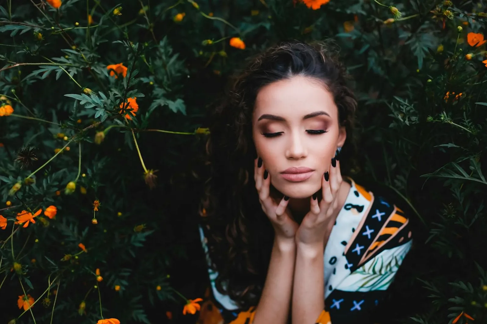 Žena sa kovrdžavom kosom i zatvorenim očima, okružena zelenilom i narandžastim cvijećem, dodiruje svoje lice rukama.