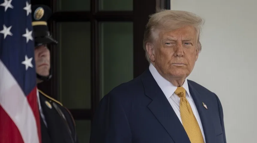 Donald Trump u tamnom odijelu i žutoj kravati, pored američke zastave i pripadnika vojne garde.