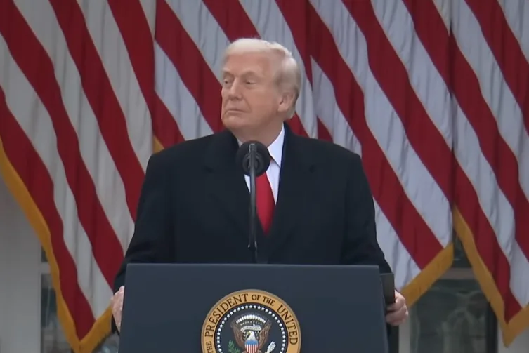 Donald Trump u crnom kaputu i crvenoj kravati govori za govornicom sa grbom Predsjedništva SAD-a, ispred zastava Sjedinjenih Američkih Država.