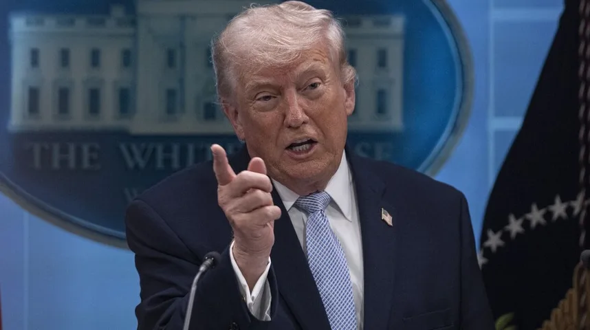 Donald Trump govori, podižući desnu ruku u znak naglašavanja, u tamnom odijelu i plavoj kravati, sa grbom Bijele kuće i američkom zastavom u pozadini.