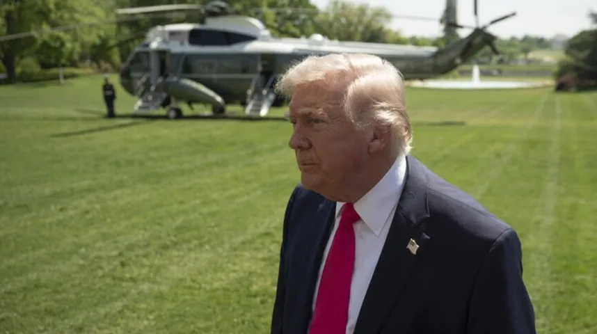 Donald Trump u odijelu s crvenom kravatom, stoji na zelenoj travi u blizini Bijele kuće s helikopterom u pozadini.