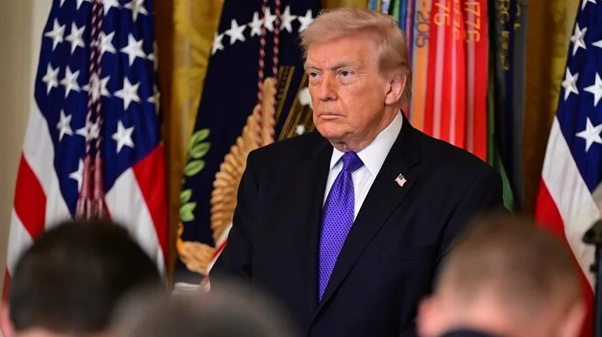 Donald Trump u tamnom odijelu i ljubičastoj kravati stoji ispred američke zastave i drugih zastava.