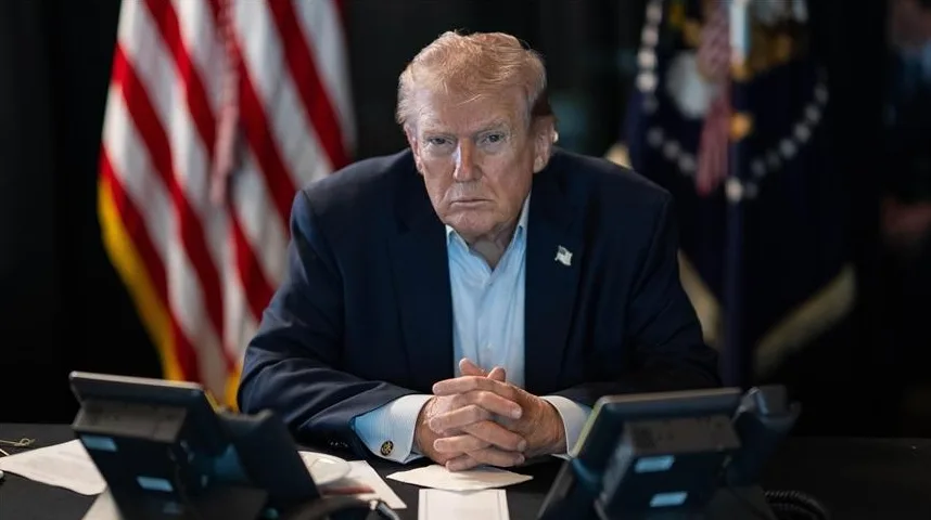 Donald Trump sjedi za stolom sa sklopljenim rukama, ispred njega su telefoni i američka zastava.