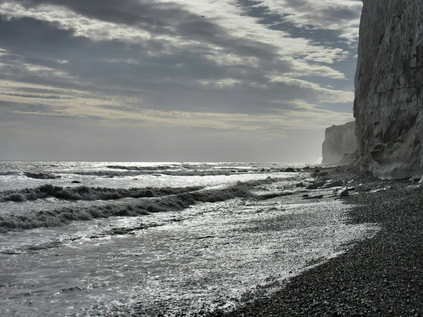 Fotografija prikazuje dramatičnu morsku obalu sa bijelim liticama na desnoj strani, tamnim šljunkom na obali i uzburkanim tamnim talasima mora koji udaraju o obalu. Nebo je oblačno sa suncem koje probija kroz oblake, stvarajući blještave odsjaje na vodi.
