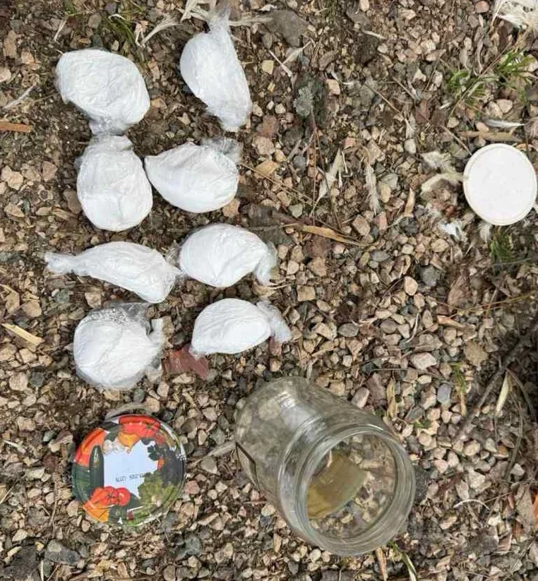 Paketi bijele materije umotani u plastiku leže na šljunkovitom tlu, pored staklene tegle i poklopca.