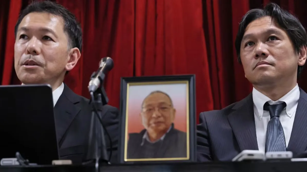 Dva muškarca u crnim odijelima stoje ispred crvene zavjese, s portretom starijeg muškarca između njih.