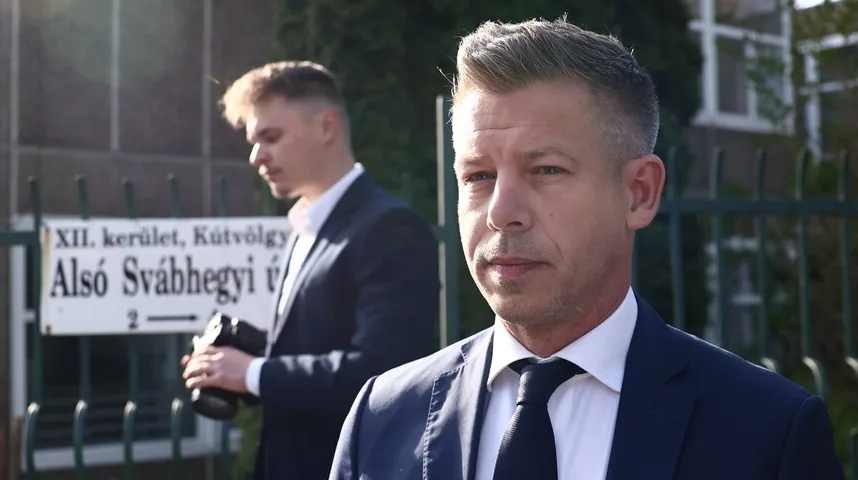 Dva muškarca u tamnim odijelima stoje ispred zgrade sa natpisnom tablom. Ispred je krupni kadar na lice jednog muškarca, dok je iza njega u odmagljenom kadru drugi muškarac koji drži fotoaparat.