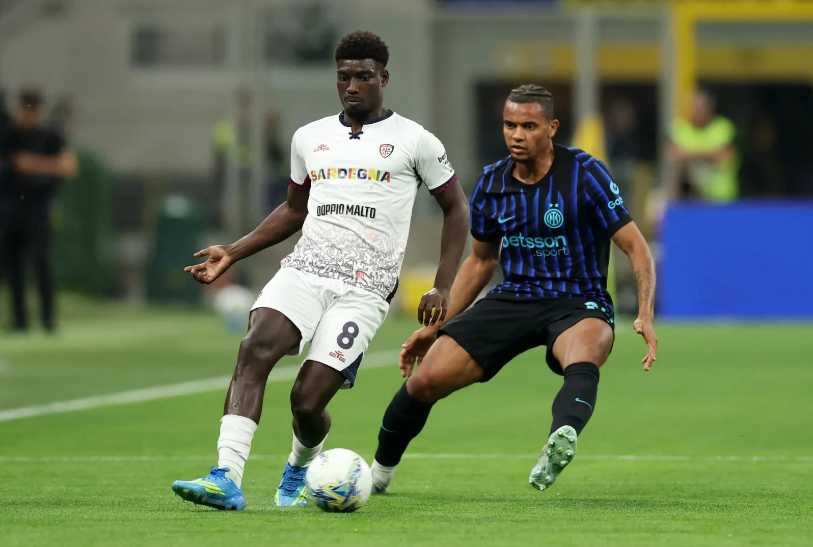 Dva nogometaša u akciji na zelenom travnjaku, jedan u bijelom dresu s brojem 8, a drugi u crnom dresu s plavim prugama.