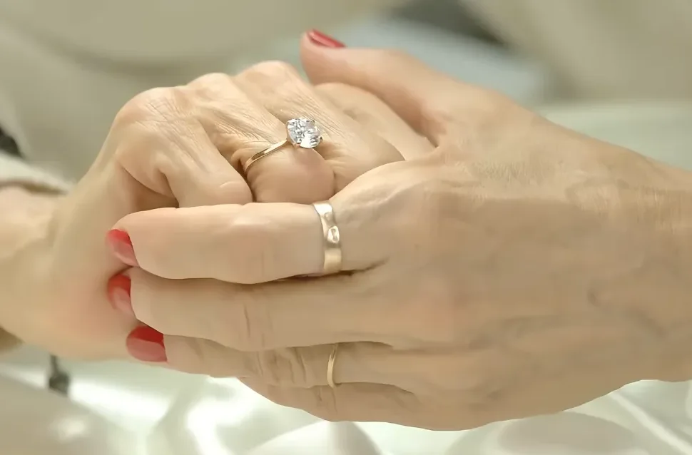 Dva para ruku, jedan iznad drugoga, sa zlatnim vjenčanim prstenjem. Na ruci koja je iznad, na prstenjaku je prsten sa dijamantom, a na prstenjaku ruke ispod, jednostavan zlatni vjenčani prsten.