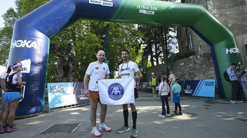 Dva muškarca u bijelim majicama i sportskim šorcevima stoje ispod velikog plavo-zelenog nadvožnjaka sa natpisima "HOKA" i "ISTRIA 100 BY UTMB". Drže zastavu sa logom "AA SPOR KLUBU" i "ANADOLU AJANSI".