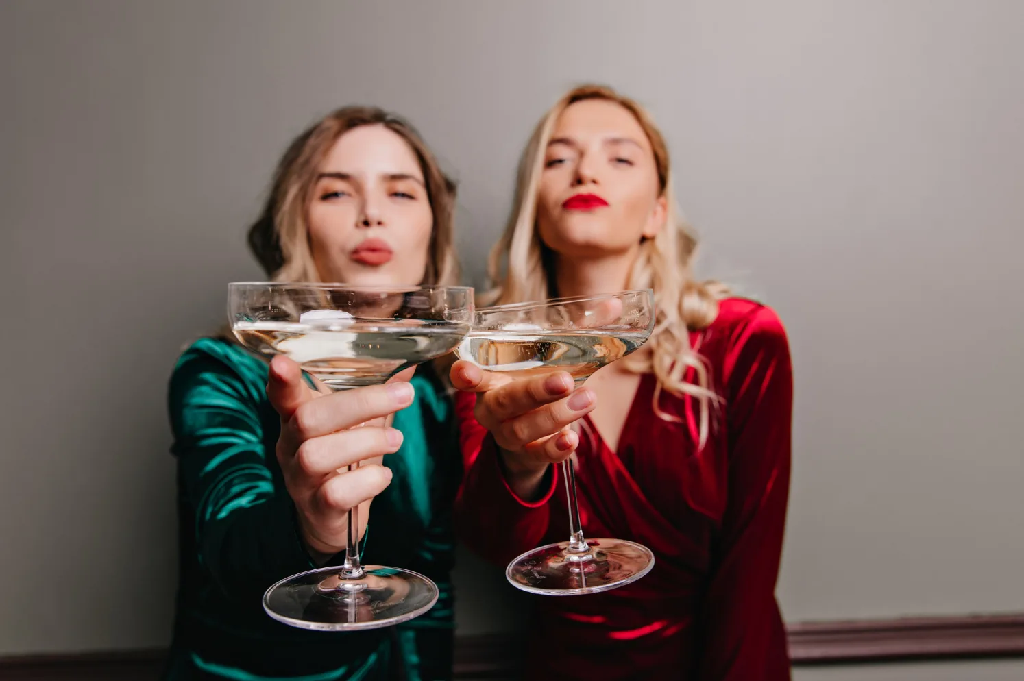 Dvije djevojke, jedna u zelenoj, druga u crvenoj haljini, drže čaše sa šampanjcem i duvaju poljupce.