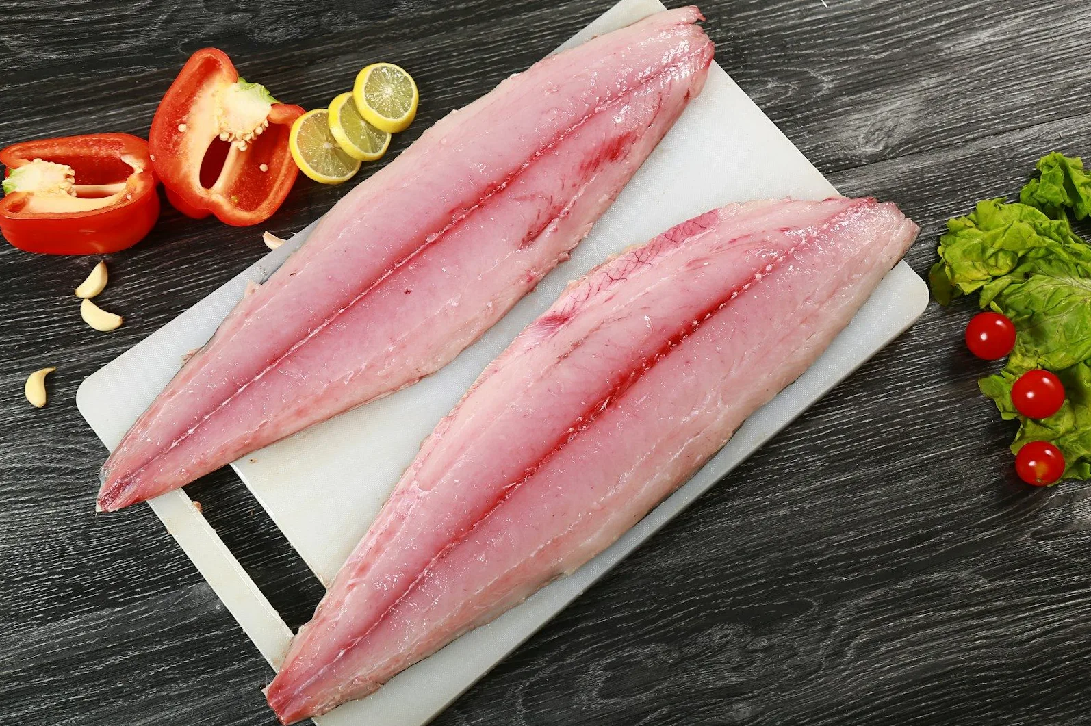 Dvije sirove, ružičaste filete ribe na bijeloj dasci za rezanje, pored crvene paprike, kriški limuna, bijelog luka, cherry paradajza i listova zelene salate, sve na tamnoj drvenoj podlozi.
