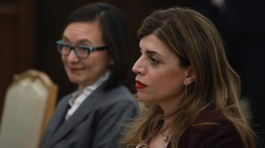 Dvije žene u odijelima sjede u zatvorenom prostoru, fokus je na desnoj ženi s tamnom kosom i crvenim ružem.