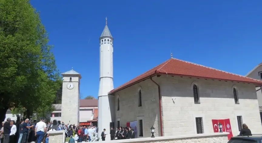 Džamija sa visokim bijelim minaretom i sat-kulom pored nje, sa grupom ljudi na događaju.
