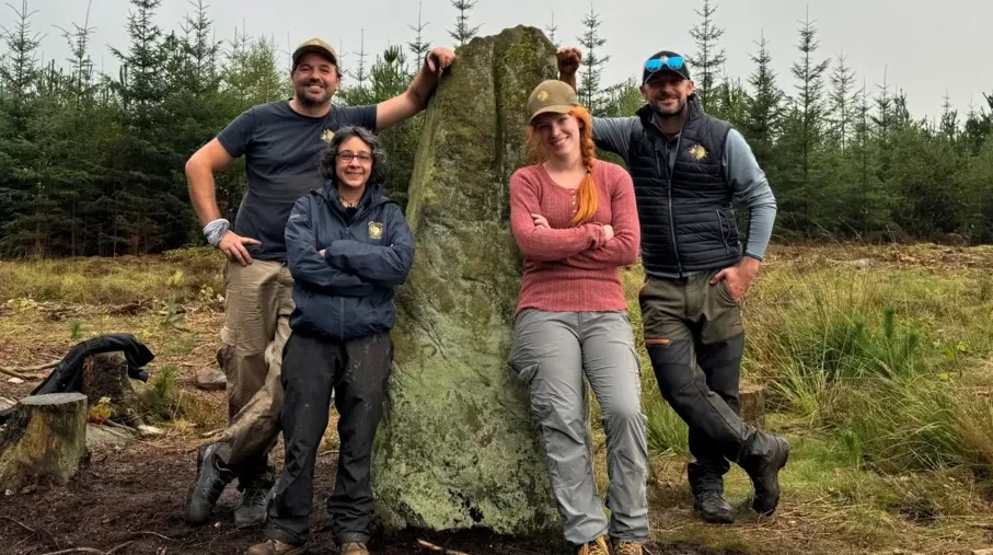 Četiri osobe stoje oko velikog, obraslog kamena u šumi.