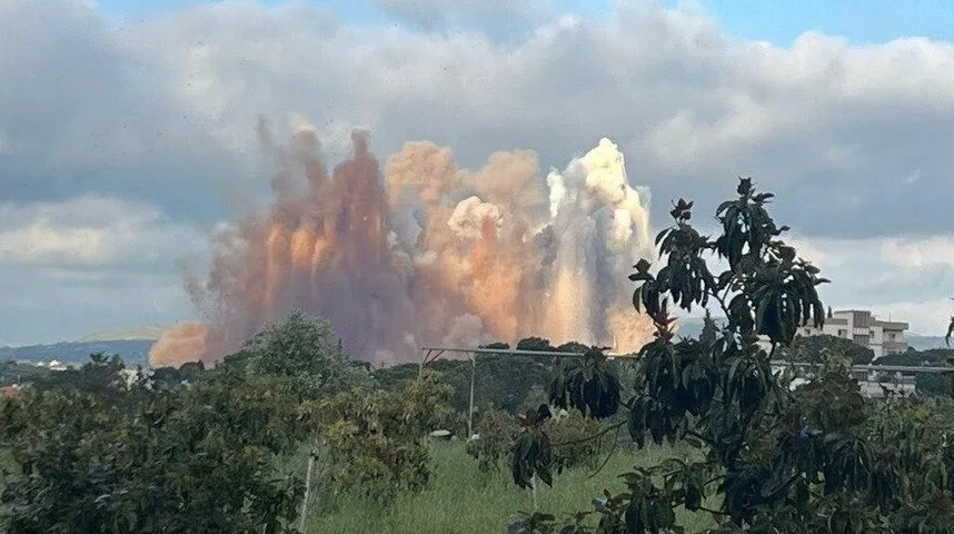 Velika eksplozija stvara masivne oblake dima i prašine koji se dižu u nebo, sa zelenilom i drvećem u prvom planu.