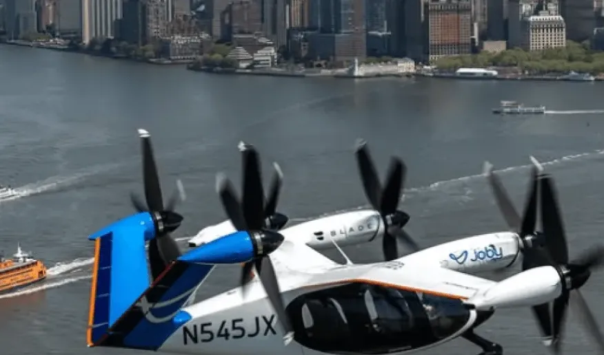 Električni eVTOL avion sa Joby logom i registarskom oznakom N545JX lebdi iznad rijeke, s gradskom panoramom i trajektom na narandžastu boju u pozadini.