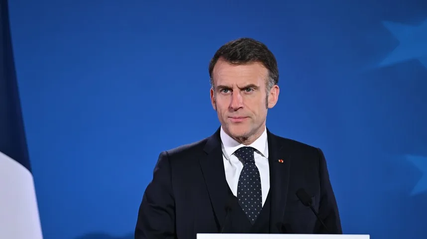 Emmanuel Macron u tamnom odijelu i s tamnom kravatom s bijelim tačkicama, obraća se javnosti pred plavom pozadinom sa zvijezdama i francuskom zastavom u lijevom uglu.