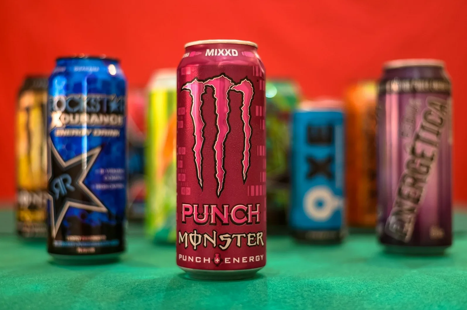 Grupa limenki energetskih pića, uključujući Monster Punch, Rockstar i druga, izložena na zelenoj površini s crvenom pozadinom.