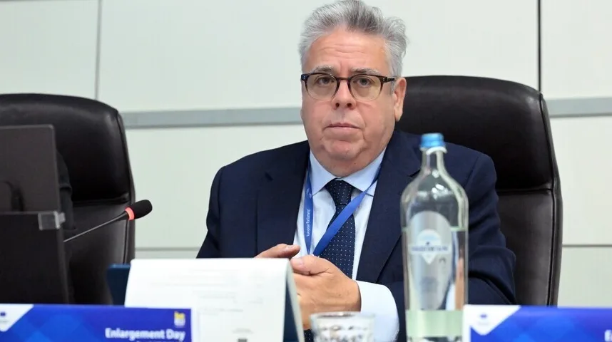 Muškarac u tamnom odijelu i plavoj kravati sa naočarima sjedi za stolom sa bocom vode i mikrofonom ispred njega.