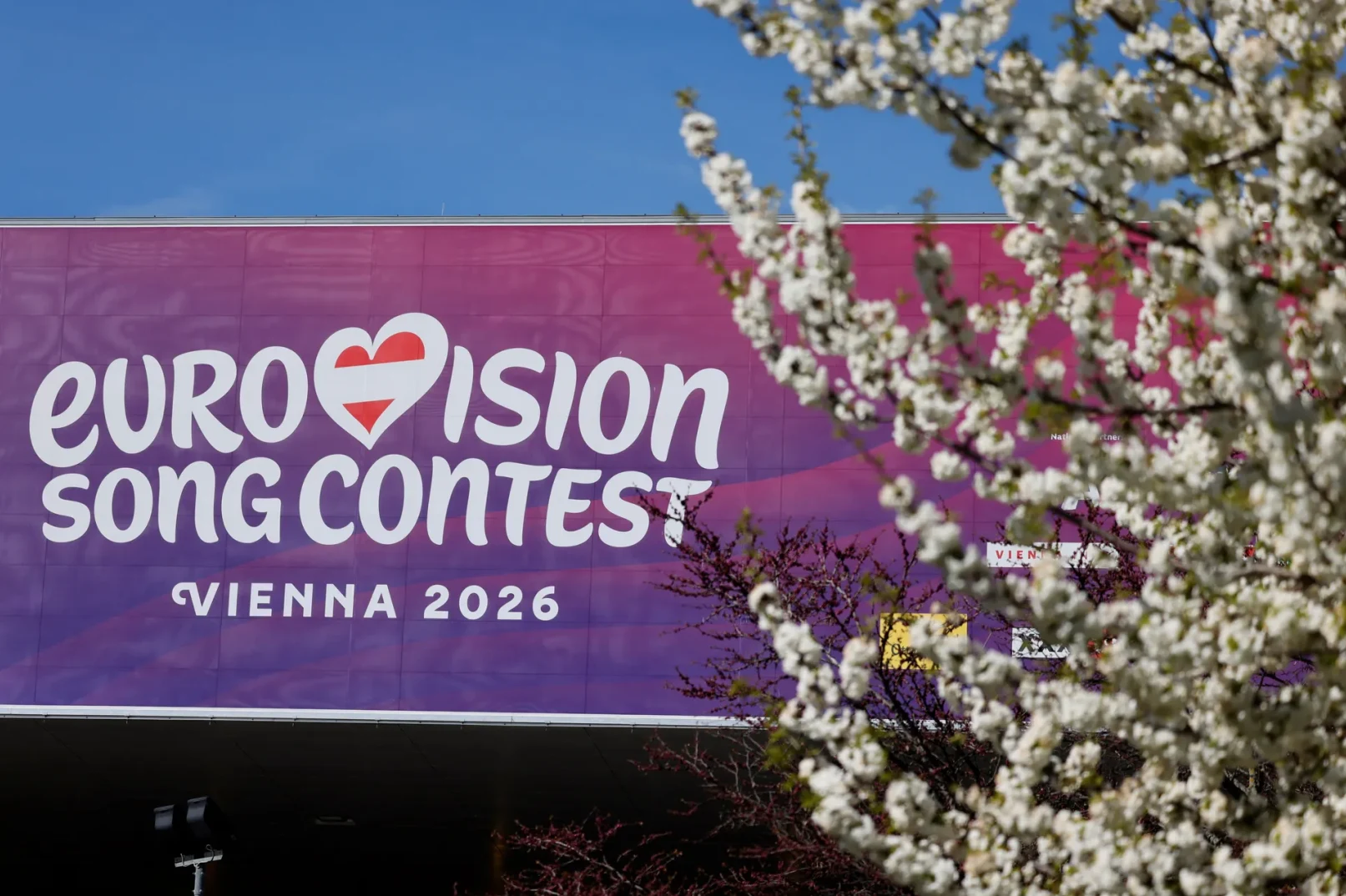Natpis "EUROVISION SONG CONTEST VIENNA 2026" na velikoj ljubičastoj pozadini, sa zastavom Austrije u obliku srca. U prvom planu su procvjetale grane.
