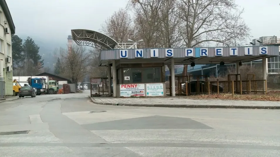 Ulaz u fabriku UNIS PRETIS sa natpisom iznad kapije, saobraćajnicom i parkiranim kamionima.