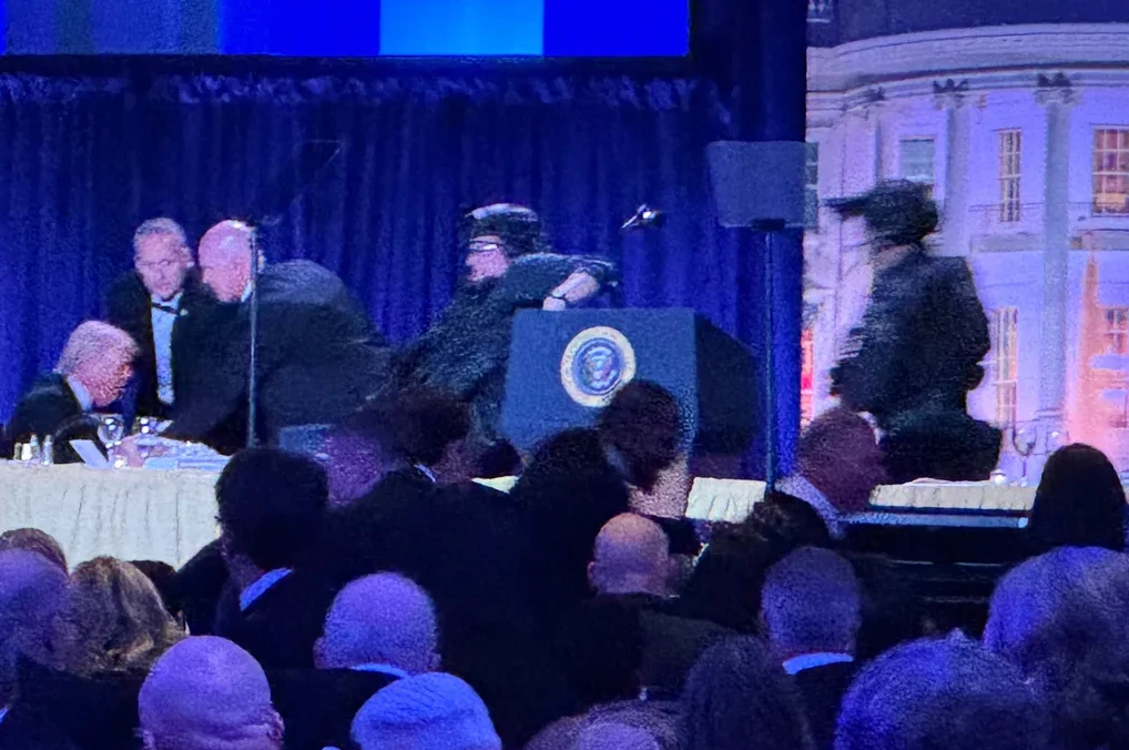 Grupa muškaraca u odijelima, od kojih neki sjede za stolom, dok je jedan u bijeloj košulji i tamnom sakou pored govornice s grbom Sjedinjenih Američkih Država. U pozadini se nazire stilizovani prikaz Bijele kuće.