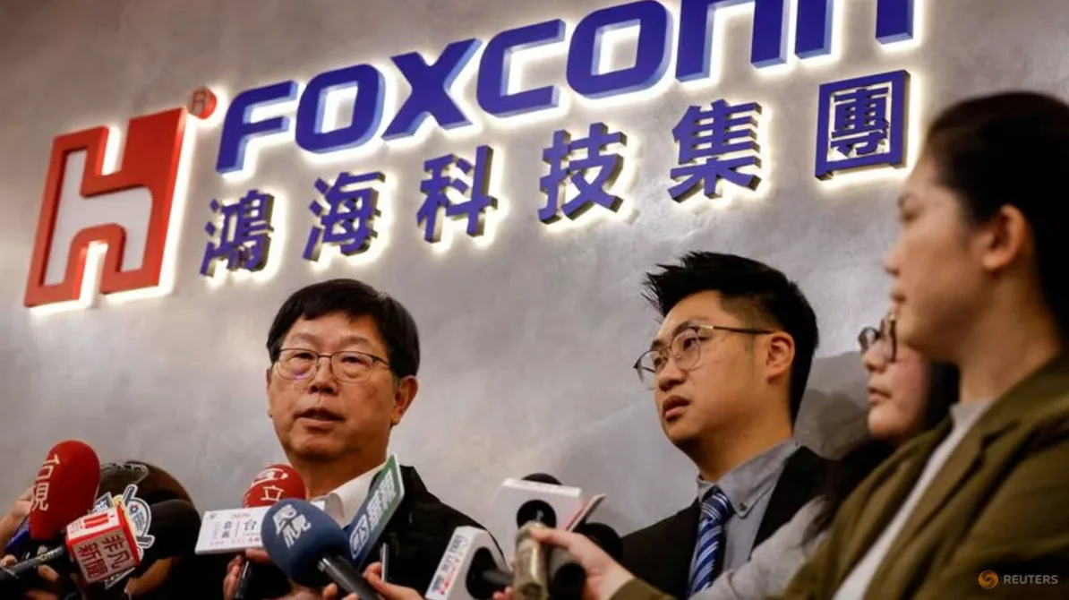 Čovjek u naočarima govori u mikrofone ispred logotipa Foxconn.