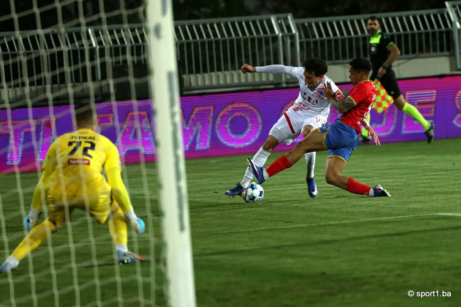 Fudbaler u bijelom dresu sa brojem 22 na leđima izbija loptu iznad fudbalera u crvenom dresu, dok golman u žutom dresu stoji u blizini gola.