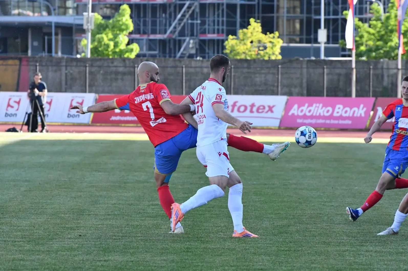 Fudbaleri u crvenim i bijelim dresovima bore se za loptu na zelenom terenu.