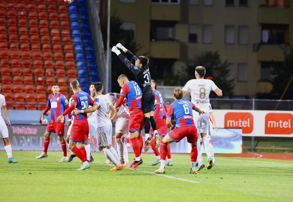 Fudbalska utakmica, golman u skoku pokušava uhvatiti loptu, igrači u crveno-plavim i bijelim dresovima.