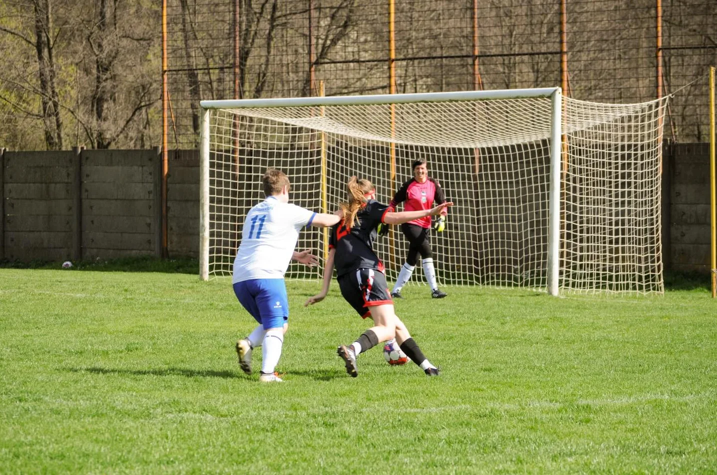 Fudbalerka u bijelom dresu sa brojem 11 šutira ka golu, dok je u blizini fudbalerka u crno-crvenom dresu i golmanica u ružičastom dresu.