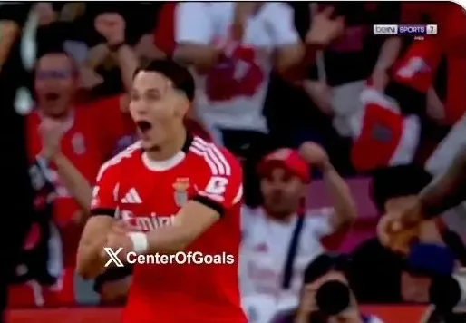 Fudbaler u crvenom dresu Benfice slavi gol sa podignutim rukama i otvorenih usta, okružen navijačima.