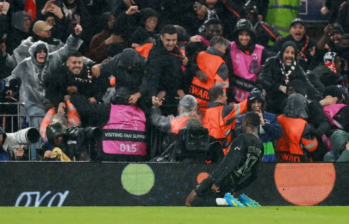 Fudbaler u crnom dresu sa brojem 17 na leđima kleči na travi i slavi gol, okružen brojnim navijačima koji ga bodre i nose kišne kabanice.