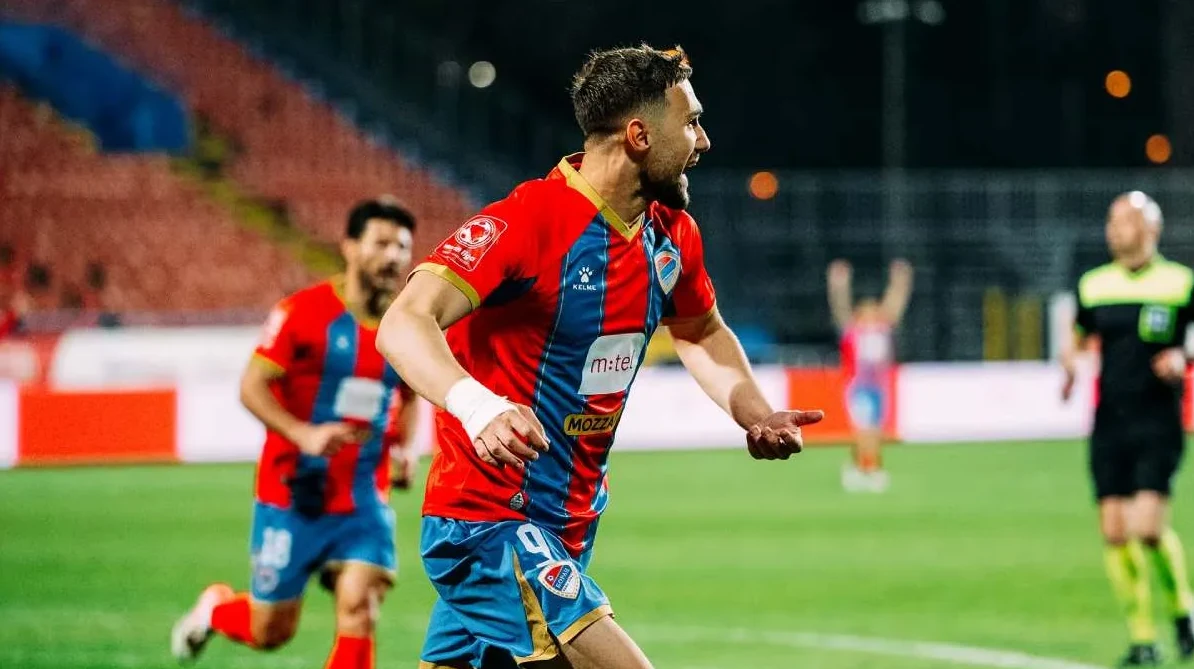 Fudbaler u crveno-plavom dresu, sa brojem 9, radosno slavi postignuti gol na fudbalskoj utakmici, sa ostalim igračima i sudijom u pozadini.