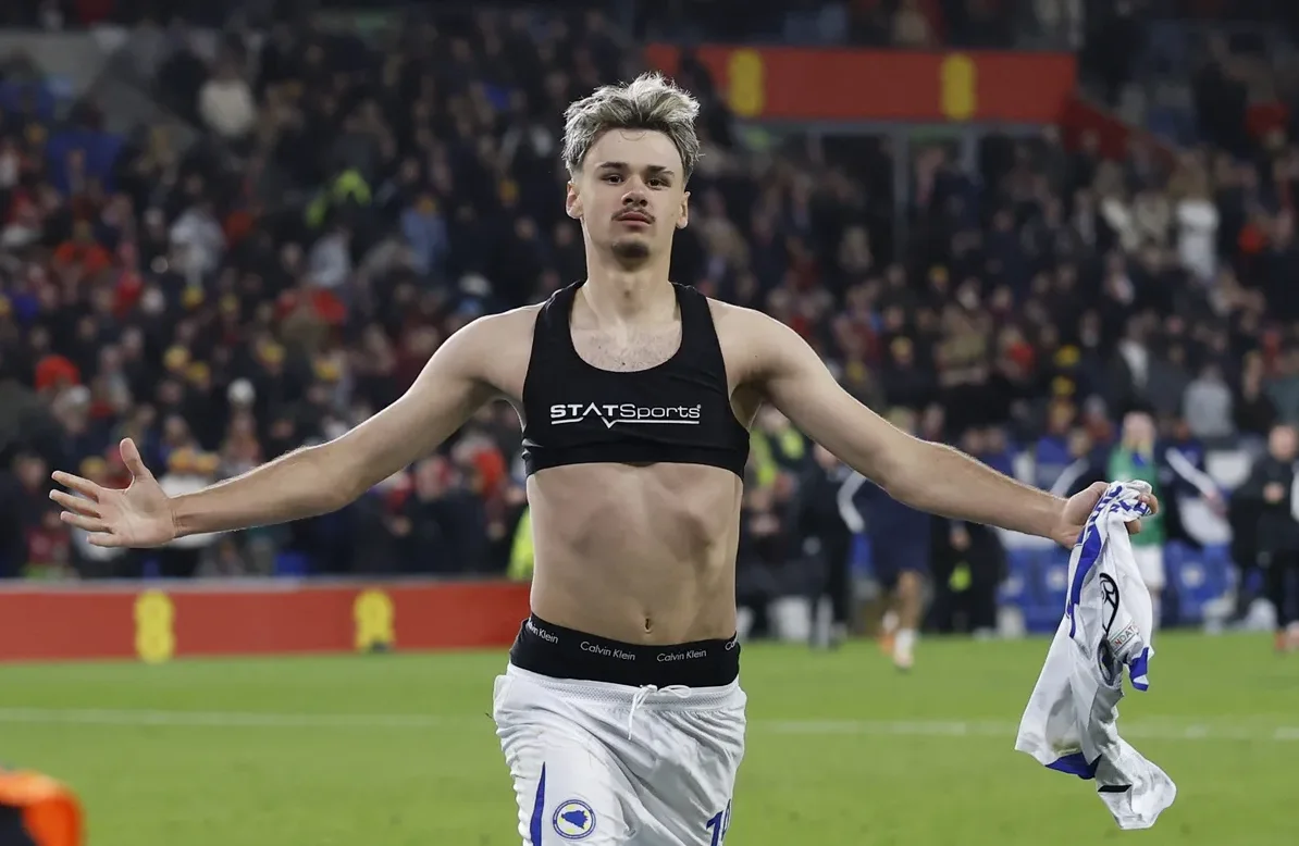 Fudbaler u crnom sportskom topu i bijelim šortsevima, sa podignutim rukama i držeći dres u ruci, slavi pobjedu na fudbalskom terenu pred publikom.