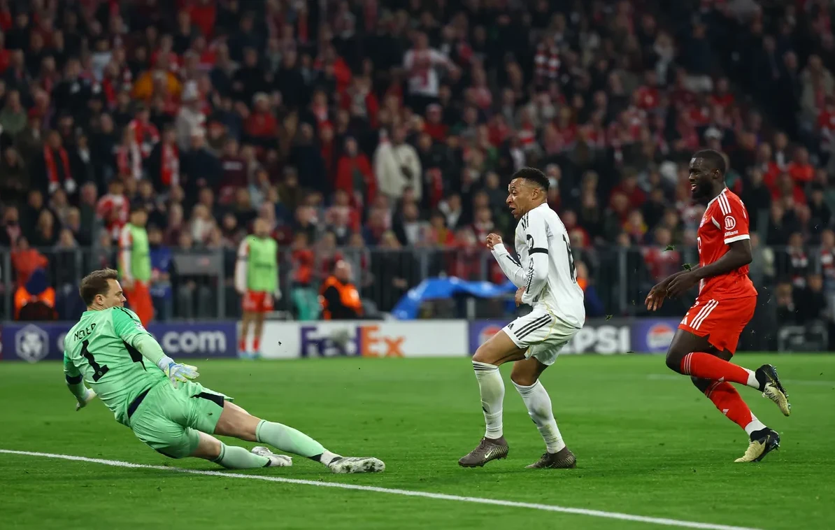 Fudbaler u bijelom dresu šutira loptu prema golmanu u zelenom dresu koji je u padu pokušava odbraniti. U pozadini se vidi nogometaš u crvenom dresu i publika na stadionu.