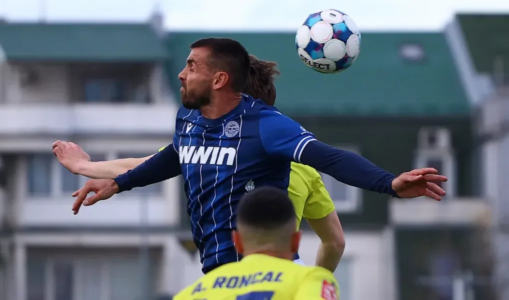 Fudbaler u plavom dresu skače i pokušava glavom udariti loptu u zraku.