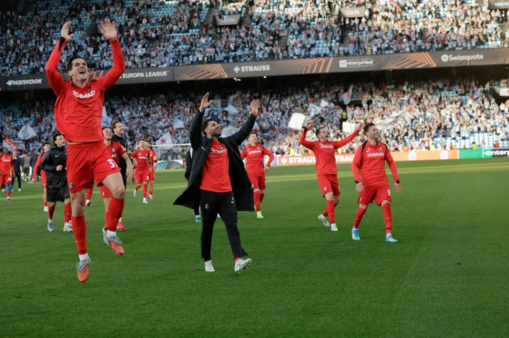 Fudbaleri u crvenim dresovima slave sa podignutim rukama na zelenom terenu stadiona, dok iza njih sjede prepuni gledaoci na tribinama.