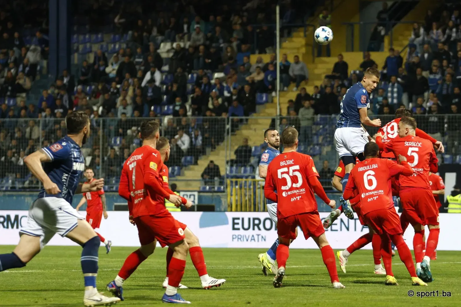 Fudbaleri u crvenim dresovima i jedan u plavom skakačiu ka lopti na stadionu